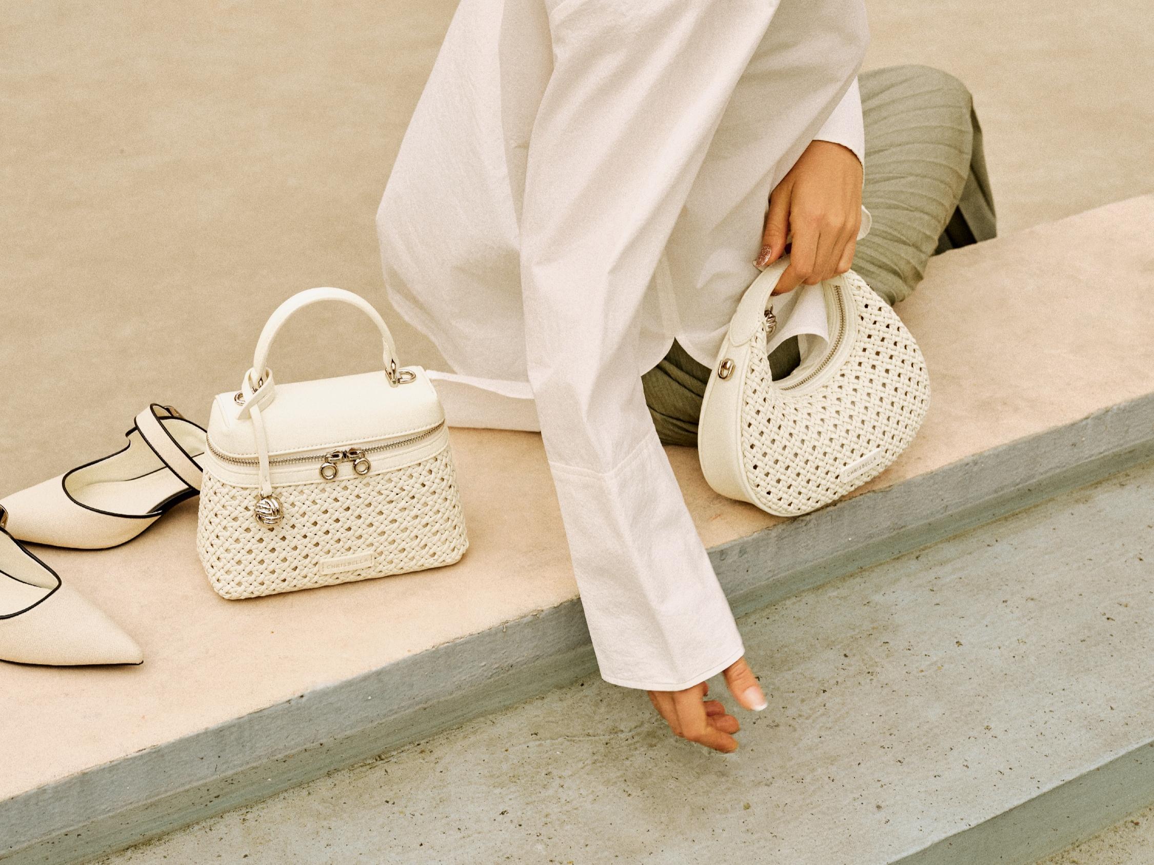 A close-up shot of a model, wearing a white shirt and sage green skirt, holding an off-white, intricately woven crescent-shaped shoulder bag. Beside her sits an off-white woven vanity-style bag with silver details and a pair of matching slingback heels. The scene has a soft, natural, aesthetic, capturing a chic, relaxed mood on a clean concrete step, perfect for illustrating spring fashion, lifestyle, and stylish, woven accessories.